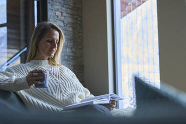 Eine Frau liest ein Buch und trinkt zu Hause Kaffee - JAHF00294