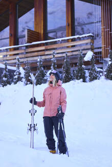 Woman wearing winter clothes holding skis in front of chalet - JAHF00304