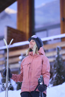 Woman with eyes closed holding skis in front of chalet - JAHF00305