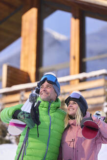 Smiling man and woman standing with skis in front of chalet - JAHF00312