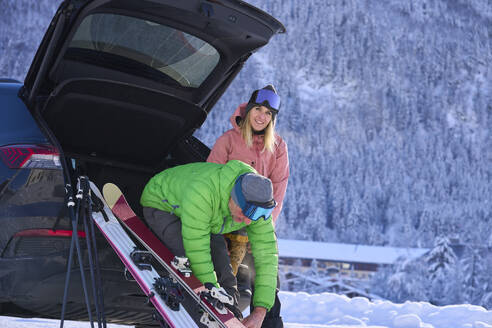 Smiling woman and man with skis sitting in car trunk - JAHF00314