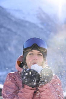 Smiling woman wearing gloves blowing on snow - JAHF00317