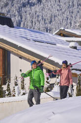 Man and woman carrying skis walking in front of chalet - JAHF00320