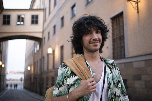 Smiling young man carrying jacket walking in alley - AXHF00345