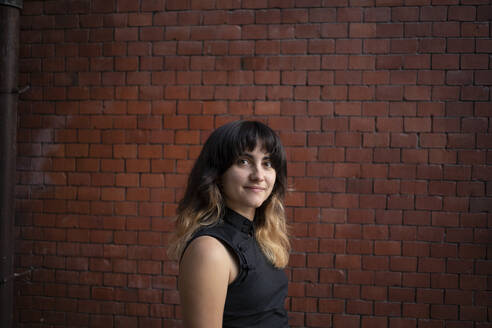 Smiling young woman standing by brick wall - AXHF00357