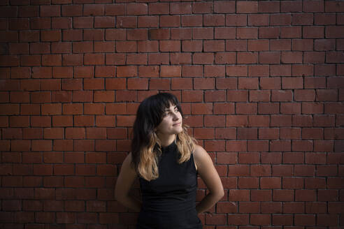 Thoughtful young woman with bangs standing in front of brick wall - AXHF00358