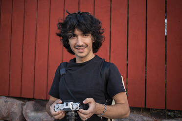 Happy man standing with camera in front of wall - AXHF00361