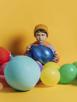 Junge sitzend mit bunten Luftballons vor gelbem Hintergrund - VSNF00579