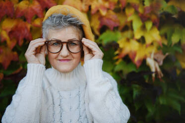 Porträt einer älteren Frau, die im Freien vor einem farbenfrohen natürlichen Herbsthintergrund steht und in die Kamera schaut. - HPIF07923