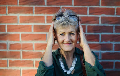Porträt einer älteren Frau, die im Freien an einer Backsteinmauer in der Stadt steht und in die Kamera schaut. - HPIF07933