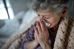 Eine kranke ältere Frau liegt zu Hause im Bett, niest und schnäuzt sich. - HPIF07970