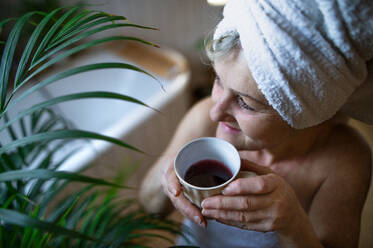 Happy senior woman wrapped in a towel in bathroom at home, sitting and resting with tea. - HPIF07981