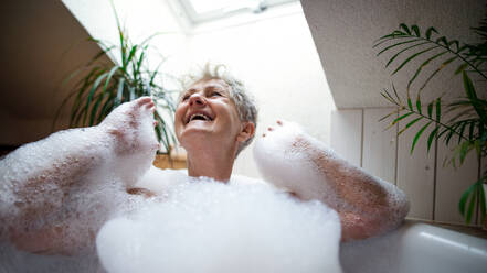 Porträt einer fröhlichen älteren Frau, die sich zu Hause in einem Schaumbad wäscht und lacht. - HPIF07992