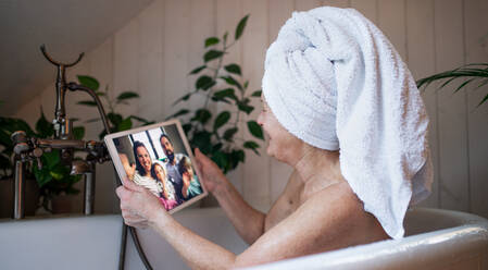 Glückliche ältere Frau mit Tablet in der Badewanne zu Hause, Videoanruf mit Tochter und Enkelkindern. - HPIF07997