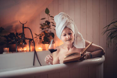 A happy senior woman reading book and drinking wine in bath tub at home. - HPIF08000