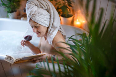 A happy senior woman reading book and drinking wine in bath tub at home. - HPIF08001