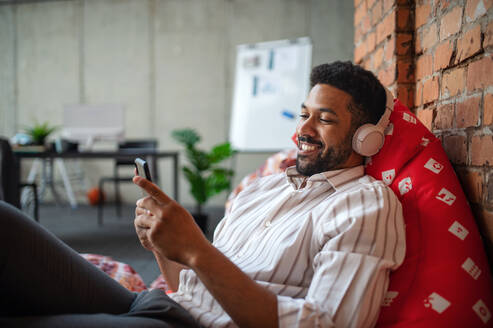 Ein fröhlicher junger Geschäftsmann mit Kopfhörern und Smartphone im Büro, der eine Pause macht und sich entspannt. - HPIF08029