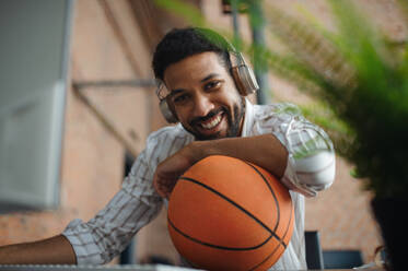 Ein fröhlicher junger Geschäftsmann mit Kopfhörern und Ball macht eine Pause im Büro und schaut in die Kamera. - HPIF08030