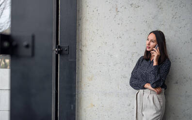 A portrait of mid adult businesswoman standing indoors in office, using smartphone. - HPIF08044