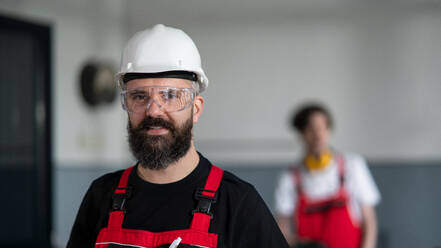 A portrait of worker with helmet and protective glasses indoors in factory looking at camera. - HPIF08127