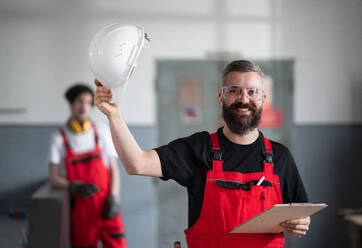 Ein Porträt eines fröhlichen Arbeiters mit Helm und Schutzbrille in einer Fabrik, der in die Kamera schaut. - HPIF08129