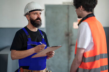Porträt von männlichen Arbeitern mit Helm, die in einer Fabrik arbeiten und sich über Probleme unterhalten. - HPIF08138