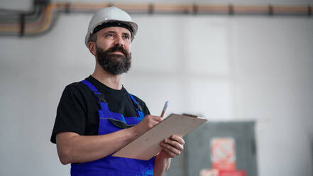 Blick aus geringer Höhe auf einen Arbeiter mit Helm in einer Fabrik, der ein Klemmbrett hält. - HPIF08140