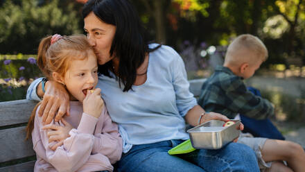 Eine glückliche ältere Frau sitzt auf einer Bank und gibt ihrer Enkelin einen Kuss und einen Snack im Freien im Park. - HPIF08291