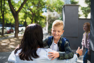 A happy schoolboy running to his grandmother waiting for him after school outdoors in street. - HPIF08299