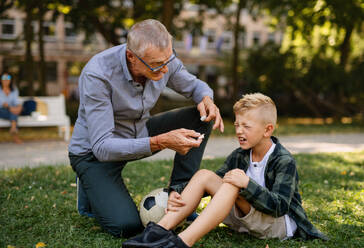 Ein kleiner Junge mit verletztem Bein weint, sein Großvater gibt ihm draußen im Park ein Pflaster. - HPIF08304