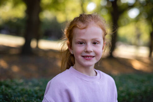 Ein glückliches kleines Mädchen mit Sommersprossen und roten Haaren schaut in die Kamera und lächelt im Freien im Park. - HPIF08310