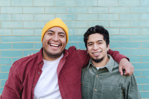 Happy man with brother standing in front of turquoise brick wall - ASGF03417