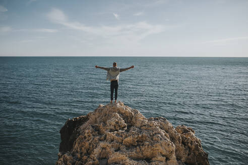 Carefree man standing with arms stretched on rock in front of sea - DMGF01048