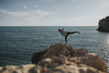 Man balancing on one leg in front of sea at rock - DMGF01051