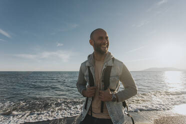 Glücklicher Mann mit Rucksack vor dem Meer stehend - DMGF01058