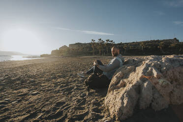 Nachdenklicher Mann sitzt am Strand bei einem Felsen - DMGF01060