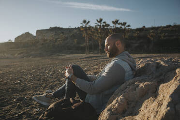 Thoughtful man sitting by rock at beach - DMGF01061