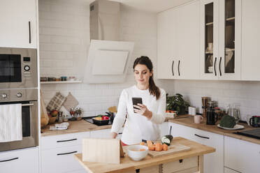 Smiling woman using mobile phone in kitchen at home - EBBF08120