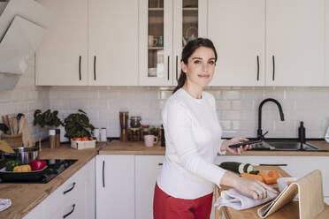 Smiling woman with tablet PC holding smart phone in kitchen at home - EBBF08121
