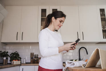 Smiling woman using smart phone in kitchen at home - EBBF08122
