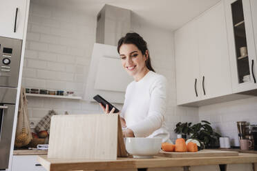 Happy woman using tablet PC holding phone in kitchen - EBBF08124