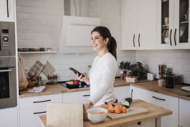 Happy woman standing with smart phone in kitchen at home - EBBF08125