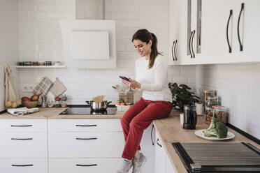 Smiling woman using smart phone sitting on counter in kitchen - EBBF08132