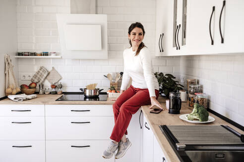 Happy woman sitting on kitchen counter at home - EBBF08133