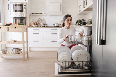 Woman arranging utensils in dishwasher at home - EBBF08139