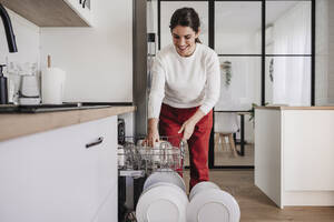 Smiling woman arranging cups in dishwasher at home - EBBF08142
