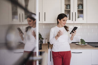 Lächelnde Frau mit Smartphone und Kaffeetasse in der Küche - EBBF08148