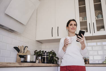 Thoughtful smiling woman with smart phone holding coffee cup in kitchen - EBBF08149