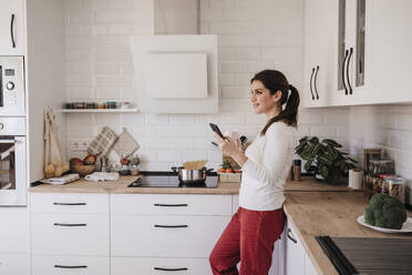 Contemplative smiling woman with smart phone in kitchen - EBBF08150