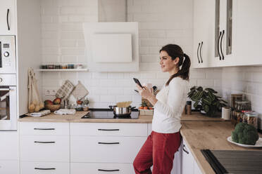 Nachdenklich lächelnde Frau mit Smartphone in der Küche - EBBF08150